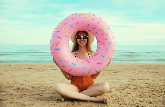 Summer vacation, tourism, happy smiling woman with swimming inflatable ring sitting on the sand on the beach on sea coast background