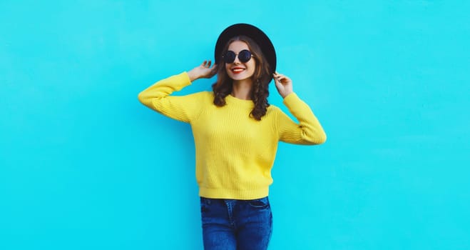 Portrait of happy stylish young woman in black round hat, yellow sweater, modern cool girl posing on blue studio background