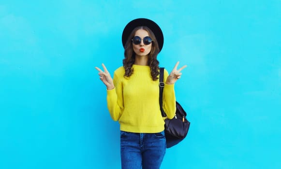 Portrait of stylish young woman in black round hat, yellow sweater with backpack, modern cool girl posing on blue studio background