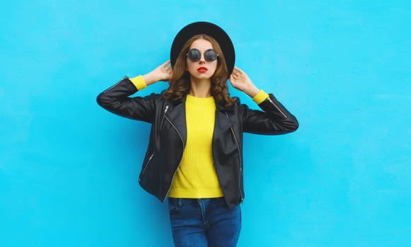 Fashion, trendy, stylish young woman in black round hat, leather jacket, yellow sweater, modern girl posing on blue studio background