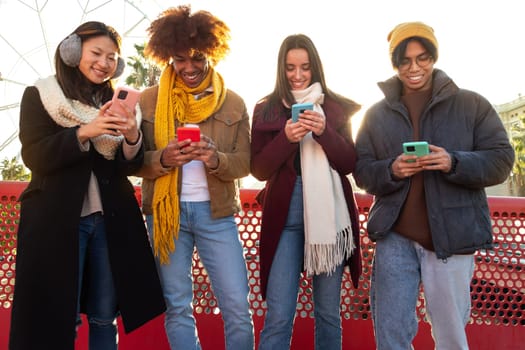 Young multiracial happy smiling people standing in a row looking at mobile phones during sunny winter day. Multiethnic friends using phones outdoors in city street.Technology and lifestyle.