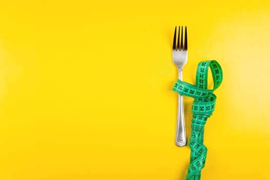 A bright green measuring tape with a fork lies on a yellow background, top view. Healthy food idea go diet for banner.