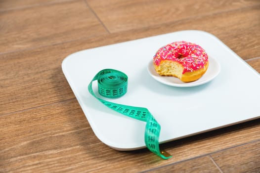 Scales for weighing with a measuring tape and a red donut next to it on the floor. Proper nutrition. Medical fasting. Slimming diet concept.