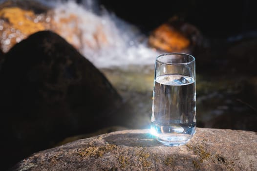 Clean water, healthy concept. Mountain mineral water in a glass goblet, on a stone with a background of a waterfall or mountain river on a sunny day.