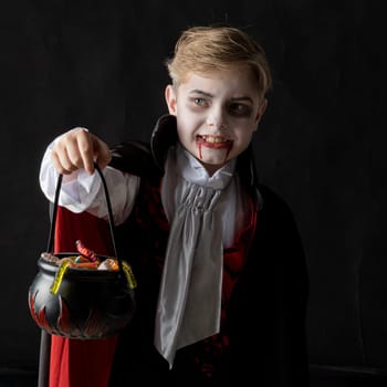 Boy with cauldron basket and worm sweets dressed as vampire for Halloween party going trick or treating. Studio portrait isolated over black background