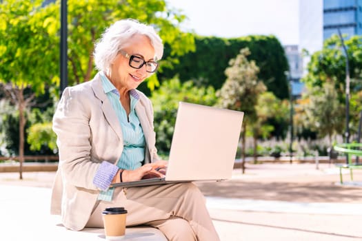 middle age woman smiling happy using laptop sitting outdoors in a city park, concept of technology and modern mature people lifestyle, copyspace for text
