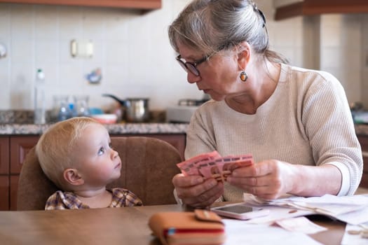 Senior woman counting money americain dollars and euro at home teaching kid financial literacy planning budget together with little child.