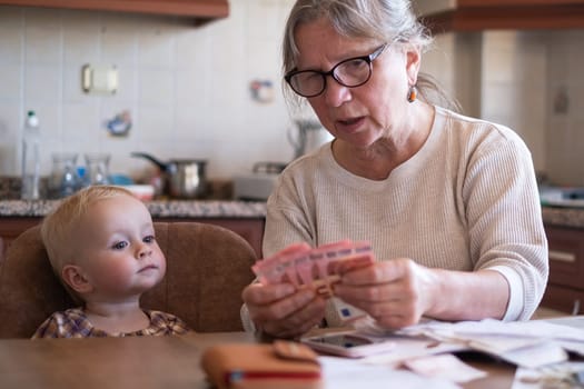 Senior woman counting money americain dollars and euro at home teaching kid financial literacy planning budget together with little child.