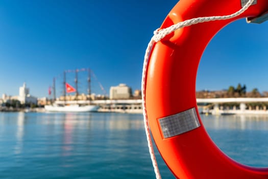 Vibrant orange lifebuoy dominates the foreground in a peaceful marina setting with docked boats, waterfront buildings, and clear blue sky download image