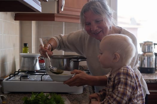 Senior old eldery aged woman grandmother for a toddler cooking soup in kitchen at home. Grandma preparing food, meal. Healthy food, vegetarian concept. Dieting and healthy lifestyle.