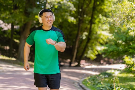 Active sportsman jogging training marathon run, listening to music in wireless headphones. Workout cardio outside in park in morning. Young guy enjoying healthy lifestyle. Japanese athlete jogger.