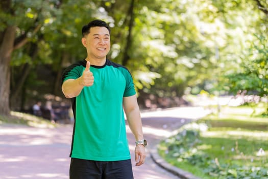 Like. Portrait athletic fit smiling runner man training looking at side happy about marathon achievement. Workout cardio in park. Japanese young guy sportsman showing thumbs up against blurred trees