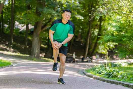 Asian runner athlete with knee pain in park. Japanese man massaging stretching, trauma injury while jogging outdoors. Fitness male guy sprain severe pain stretch pull. Leg muscle cramp calf sport.