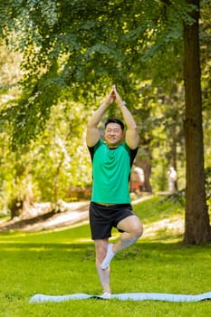 Calm, muscular Japanese sportsman balances on one leg on mat in summer park. Fit Asian guy yogi is training for well-being. Male stands in Tree Pose, stretching his leg in nature, focusing on balance.