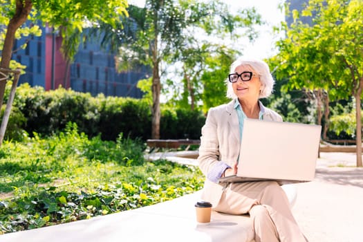 middle age woman smiling happy using laptop sitting outdoors in a city park, concept of technology and modern mature people lifestyle, copyspace for text