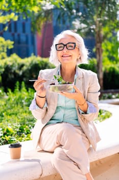 middle age entrepreneur woman smiling happy while eating a takeaway salad sitting outdoors in a city park, concept of healthy food and active lifestyle in adulthood
