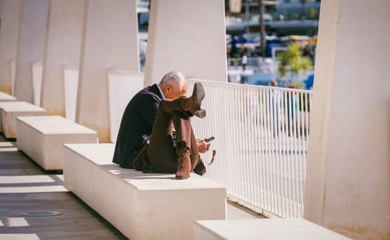 Elderly person resting on a bench near a marina, portraying contemplation or distress, surrounded by architectural elements in daylight. High quality photo