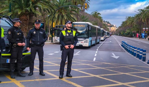 three police officers standing near a roadside. They are wearing dark uniforms with reflective elements and insignia typically seen on law enforcement apparel. One of the officers appears to be holding a notepad and pen, potentially for recording information. In the background, there is a long line of buses parked along the curb of a wide street. This, combined with the presence of the police officers, might imply a coordinated event or a situation that requires police presence and control of traffic flow. The street is lined with palm trees