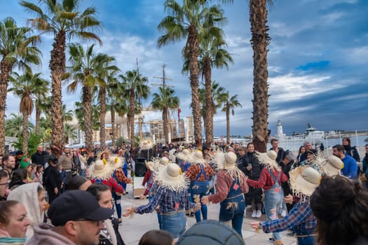 outdoor scene with a group of people engaged in what appears to be a dance performance or social dance gathering. They are wearing large straw hats, which are often associated with traditional or folkloric dance styles from various cultures. The participants are holding hands and appear to be performing a dance in a circular formation. Spectators surround the dancers, watching the performance