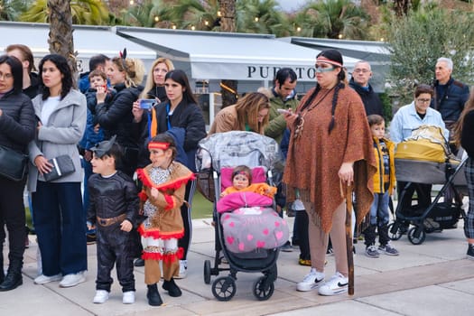 group of people outdoors, possibly at an event or gathering. In the foreground, there are two children dressed in costumes, with a boy on the left dressed as a pirate and a girl on the right dressed in a Native American-themed outfit. There is a baby in a pink stroller between them, and a woman who appears to be holding onto the stroller and watching something in the distance