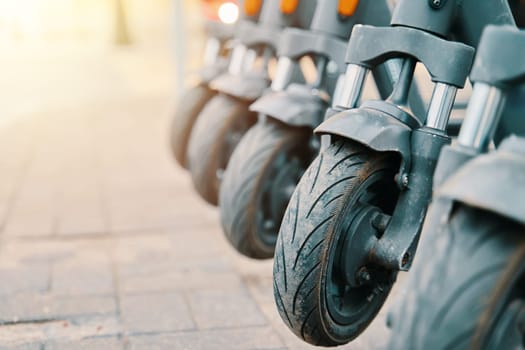 A line of scooters are parked on the sidewalk, resembling a sculptural row of art pieces made from metal, wood, and automotive tire materials