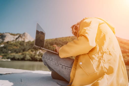 A young woman sits on a lakeside, wearing a yellow raincoat and jeans, working on a laptop.