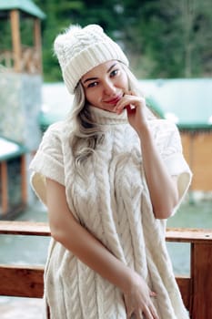 Portrait of a beautiful girl in a white hat and sweater on the street on the balcony. Warm winter clothes.