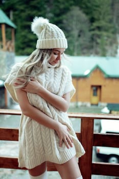 A sexy girl in a white sweater and stockings on the balcony admires the beauty of nature. wool clothes and a hat on a beautiful blonde.