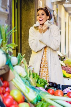 Positive young female greengrocer, in casual warm clothes with bunch of fresh fruits and vegetables looking at camera while standing and making punch fist in daylight