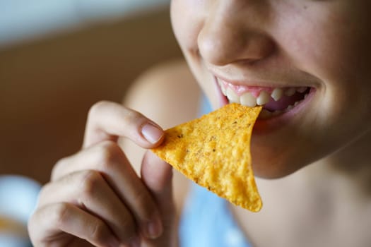 Closeup of crop anonymous young girl smiling while eating spicy Mexican tortilla chip at home