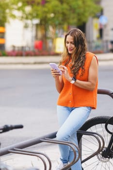 Vertical photo of a casual blonde beauty latin woman using phone sitting on public renting bike station