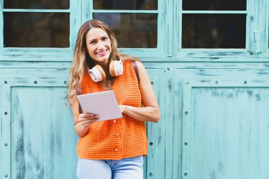 Latin woman with headphones using digital tablet standing in the city leaning on a blue wooden entrance