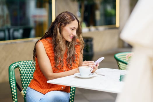 Latin smiling woman using mobile phone sitting at sidewalk cafeteria
