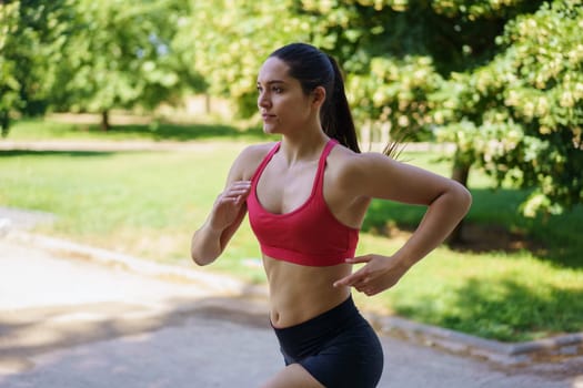 In a sunny park, a determined young woman in red sports bra and black shorts exemplifies an active, athletic lifestyle filled with energy and focus, showcasing strength and endurance