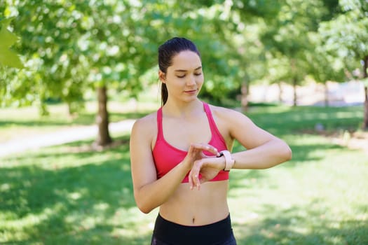 In a vibrant green park, a dedicated young woman in a pink sports bra focuses on her health and fitness routine as she checks her fitness tracker. She is committed to staying active outdoors