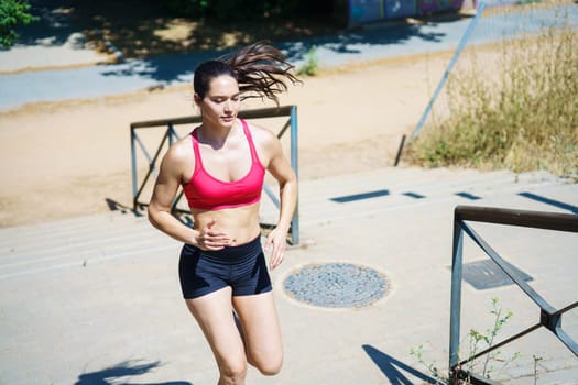 A determined female athlete in sportswear is focused on fitness, jogging up stairs in the park. She dedicates herself to outdoor exercise, maintaining strength and health under the sun
