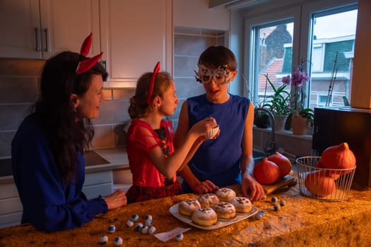 Mother, daughter and son in Halloween costumes draw skulls with cocoa powder and powdered sugar on sweet buns in a darkened room, baking together a traditional dessert for the holiday