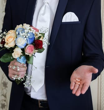 The groom in a dark suit, white shirt and tie, holding in one hand a bouquet of the bride, in the other hand two gold wedding rings.