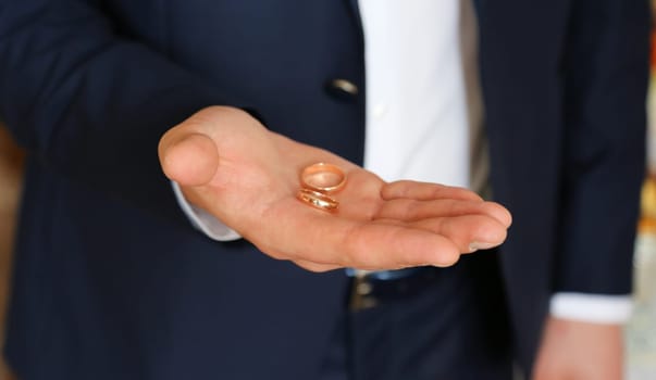 The groom in a blue suit and white shirt holds two gold wedding rings in the palm of his hand.