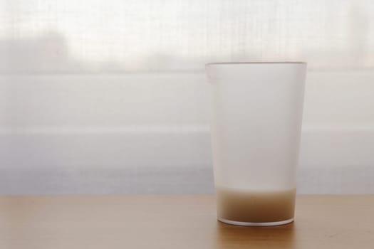 A large glass with an unfinished drink on the table against the background of a white window with a curtain.