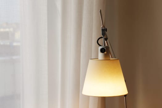 A table lamp with a lampshade glowing yellow against the background of curtains and a window. Movable table lamp with a burning light bulb.