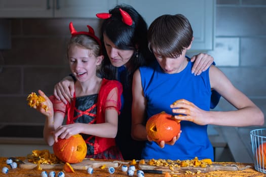 Mother, daughter and son in Halloween costumes carve eyes and teeth on pumpkins in a darkened room, traditional autumn decorations for the holiday