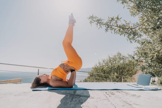 A young woman in orange sportswear practices yoga in a supported headstand pose outside on a patio with a laptop and a view of the ocean.