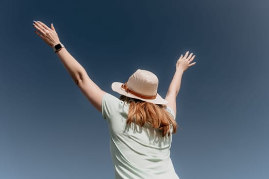 A woman wearing a straw hat and a light green shirt raises her arms to the blue sky.
