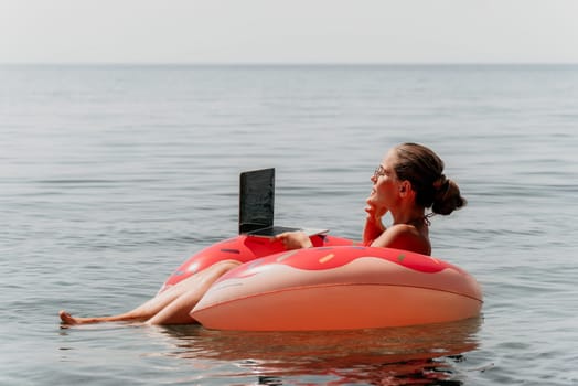 Woman freelancer works on laptop swimming in sea on pink inflatable ring. Pretty lady typing on computer while floating in the sea on inflatable donut at sunset. Freelance, remote work on vacation