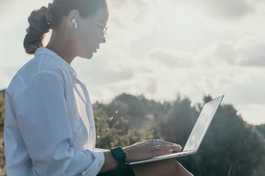 Digital nomad, woman in the hat, a business woman with a laptop sits on the rocks by the sea during sunset, makes a business transaction online from a distance. Freelance, remote work on vacation.