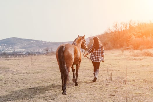 A woman in a plaid shirt walks with a brown horse in a field at sunset. The sun is setting, creating a warm glow.