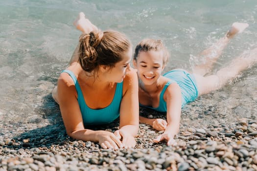 Two women in blue swimsuits are lying on a pebble beach by the water. The woman in the foreground is looking at the woman behind her, who is smiling.