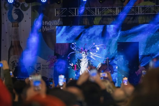 festive event or performance. In the forefront, there are many people holding up their phones, presumably taking pictures or recording the event. In the background, there's a brightly illuminated stage with blue hues and a figure that appears to be part of a performance or show