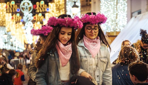 two girl walking through what appears to be an outdoor festive environment, such as a Christmas market or similar holiday event. They are wearing matching pink feathered headbands and pink scarves